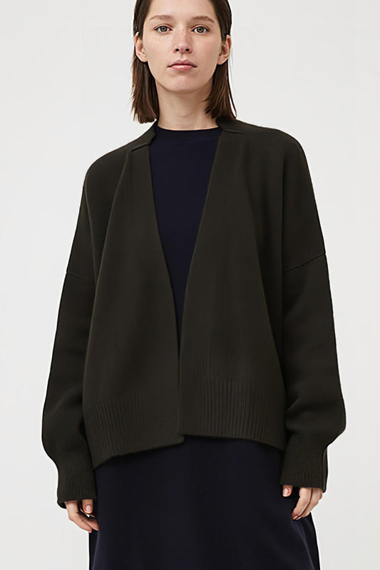 This image features the Aire Cardigan, a luxurious, chunky open cardigan made from pure cashmere. The cardigan is displayed on a model, showcasing its sleek forest color and stylish design elements.