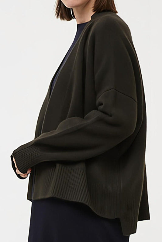 This image features the Aire Cardigan, a luxurious, chunky open cardigan made from pure cashmere. The cardigan is displayed on a model, showcasing its sleek forest color and stylish design elements.