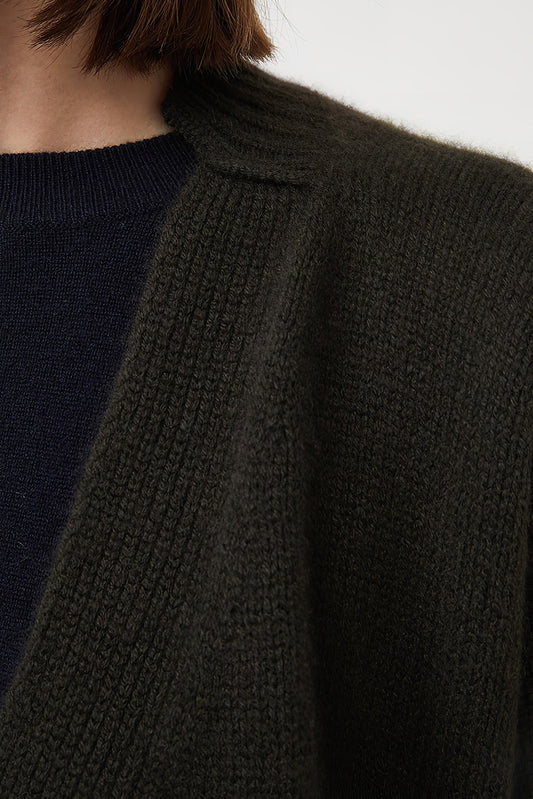 This image features the Aire Cardigan, a luxurious, chunky open cardigan made from pure cashmere. The cardigan is displayed on a model, showcasing its sleek forest color and stylish design elements.