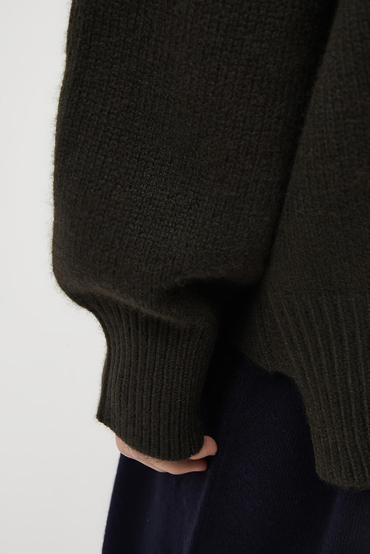 This image features the Aire Cardigan, a luxurious, chunky open cardigan made from pure cashmere. The cardigan is displayed on a model, showcasing its sleek forest color and stylish design elements.
